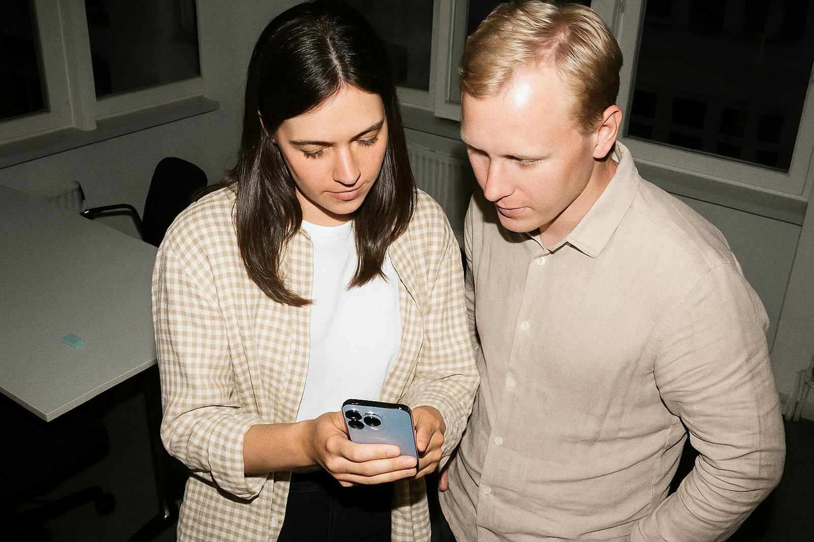 Two people in casual attire closely examining a smartphone in an office-like setting, with a focus on collaboration.